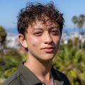 Head-and-shoulders portrait of a person with short curly hair against a blurred outdoor background.